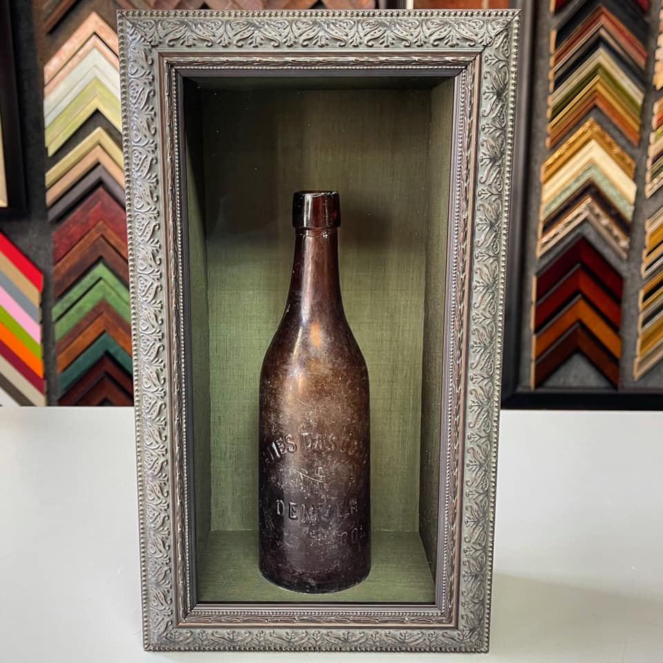 Antique glass bottle displayed in a custom shadowbox with green fabric backing and an ornate silver frame